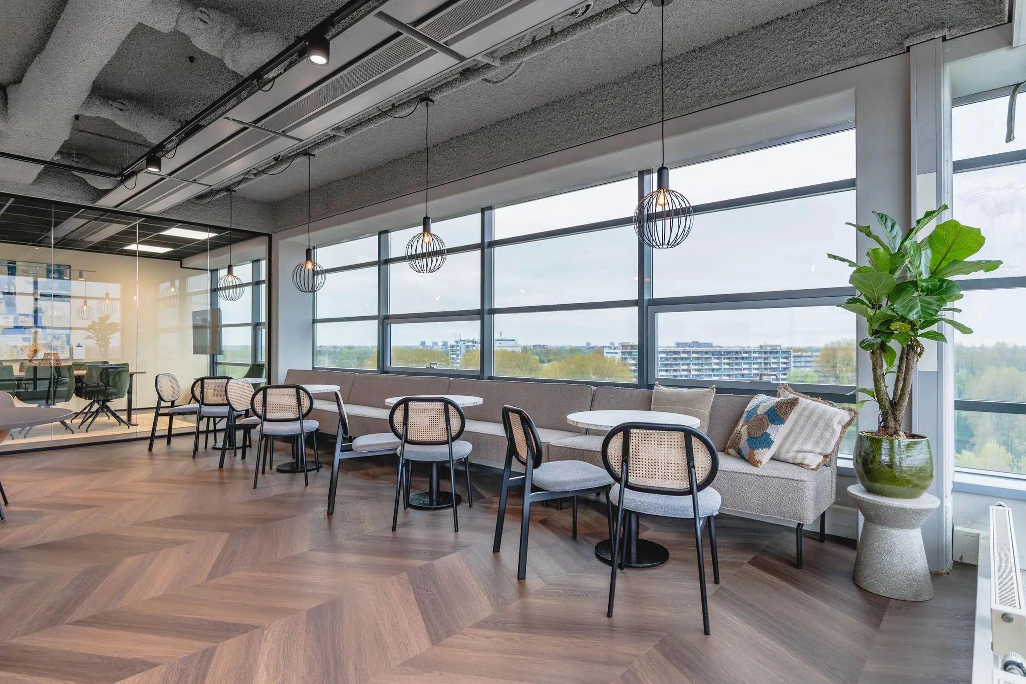 Moderne zithoek in een kantoor aan de Joop Geesinkweg met ronde tafels, designstoelen, grote ramen en uitzicht op een woonwijk.